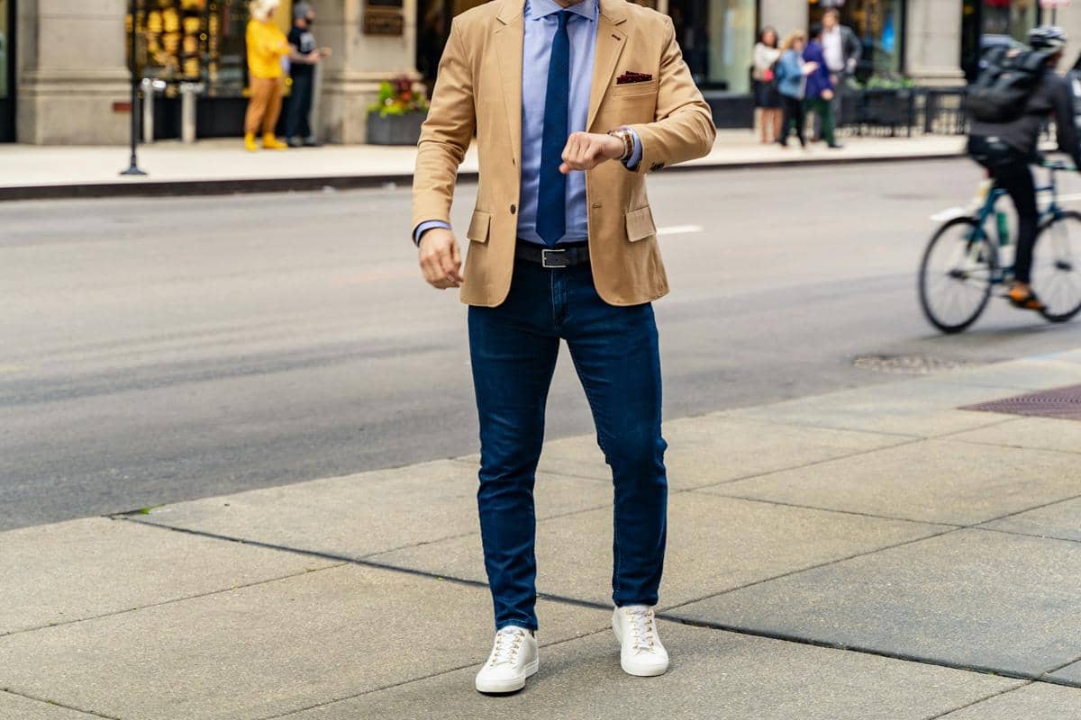 Pria mengenakan jas cokelat muda, kemeja biru, dasi biru tua, celana jeans, dan sepatu kets putih di trotoar kota.