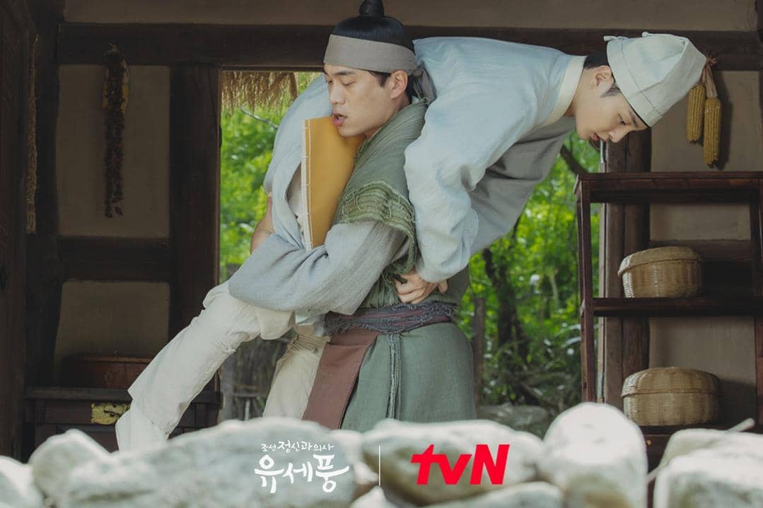 Dua pria mengenakan pakaian tradisional Joseon, salah satunya menggendong yang lain di dalam rumah kayu dengan latar pepohonan hijau.