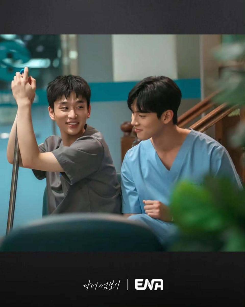 Dua pria mengenakan seragam medis sedang duduk dan tersenyum di area rumah sakit dalam cuplikan drama Doctor On The Edge.