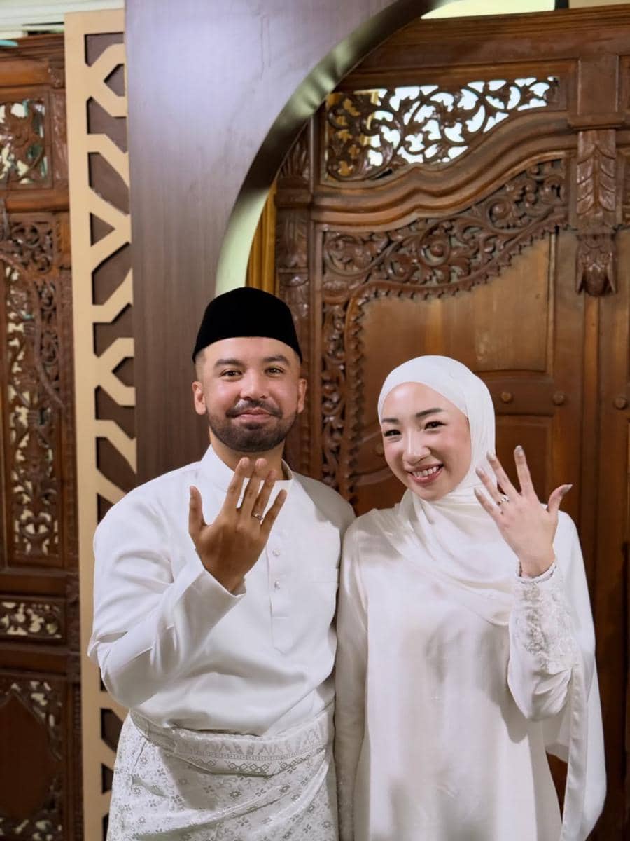 Sepasang pengantin mengenakan busana putih tradisional menunjukkan cincin pernikahan mereka di depan ukiran kayu di KUA.