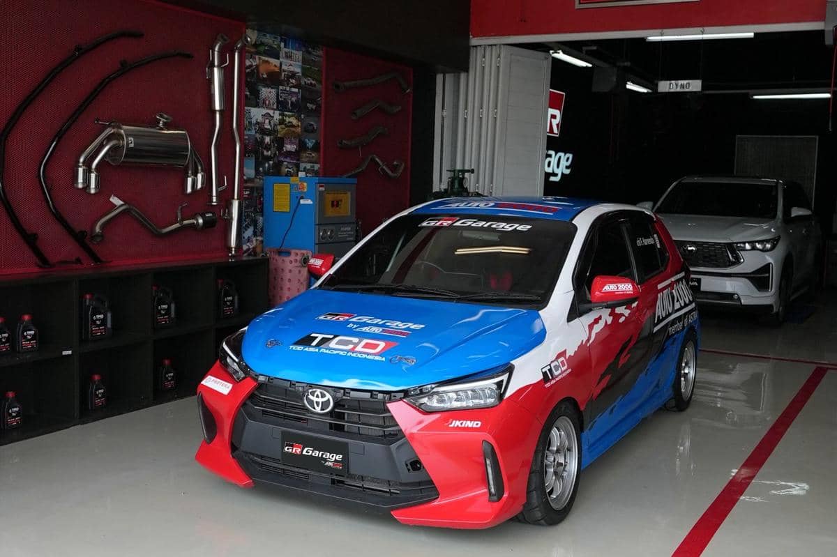 Toyota Agya berwarna merah, putih, dan biru dengan livery GR Garage berada di bengkel Auto2000 yang dilengkapi peralatan otomotif.