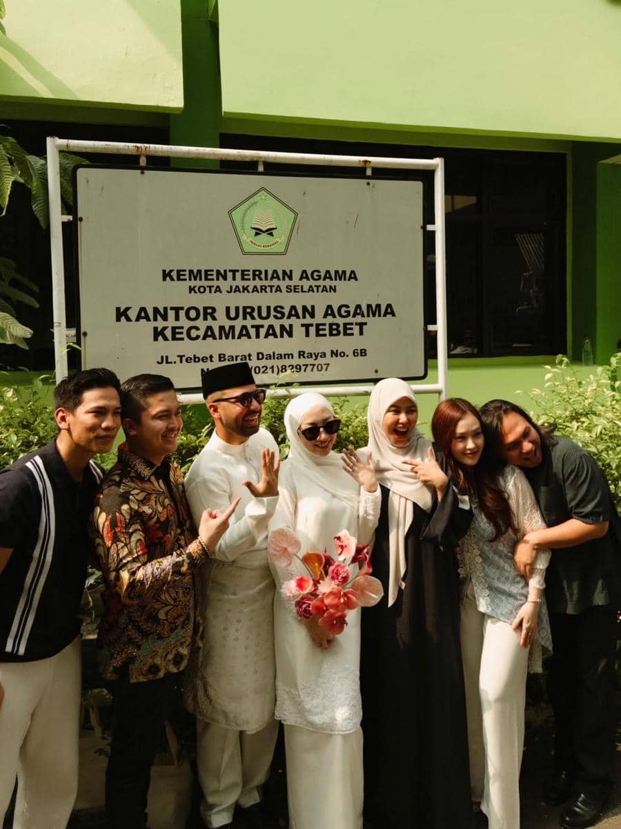Pasangan pengantin berbusana putih berpose bersama teman-teman di depan papan Kantor Urusan Agama Kecamatan Tebet, Jakarta Selatan.