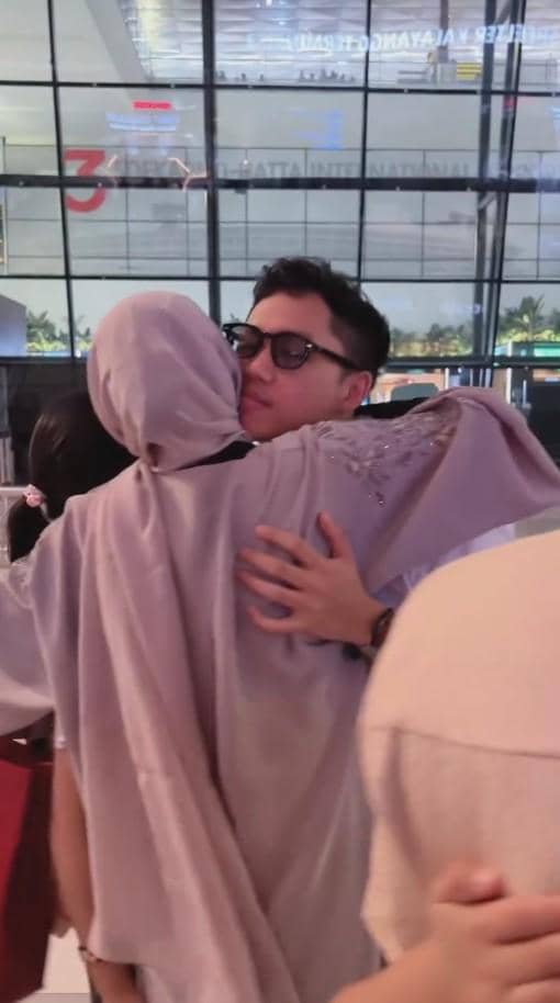 Seorang pria mengenakan kacamata memeluk wanita berhijab di area kedatangan bandara dengan suasana hangat dan emosional.
