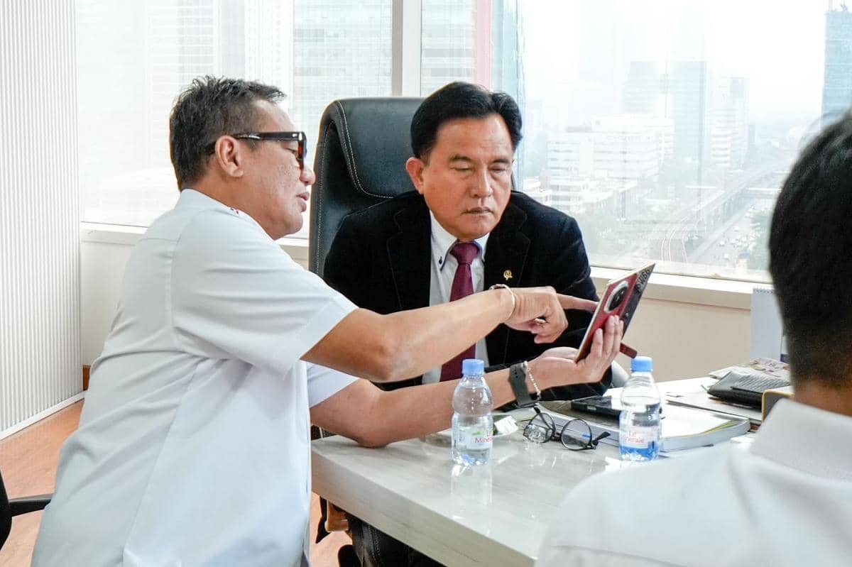 Dua pria berdiskusi di ruang kantor dengan latar gedung tinggi, salah satunya menunjukkan sesuatu di layar ponsel.