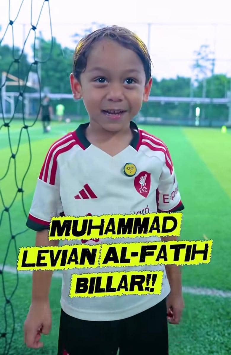 Seorang anak kecil mengenakan jersey sepak bola berwarna putih dengan garis merah berdiri di lapangan hijau dekat jaring gawang.