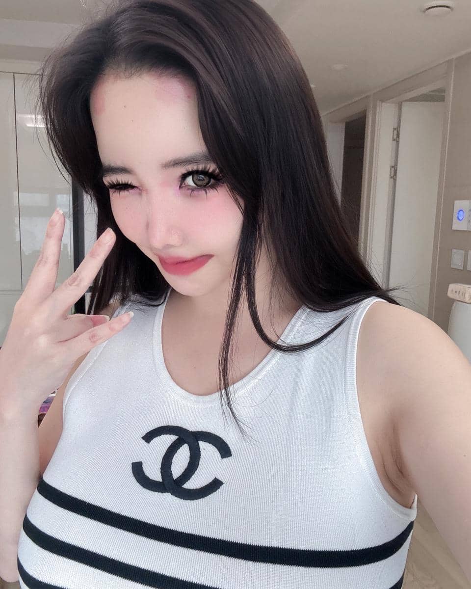 Seorang wanita berpose dengan mata terpejam dan tangan membentuk simbol jari, mengenakan atasan putih bermotif logo Chanel di dalam ruangan.