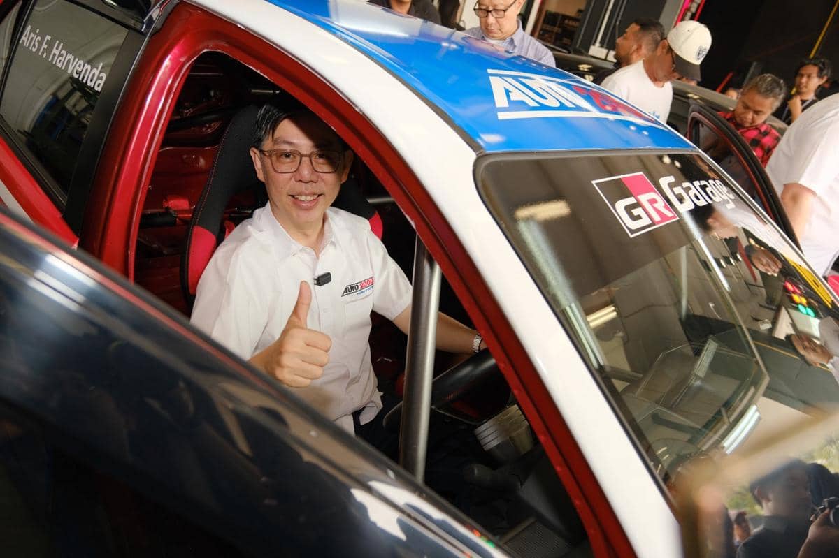 Seorang pria mengenakan kemeja putih Auto2000 duduk di dalam mobil balap dengan jempol terangkat, dikelilingi beberapa orang di bengkel.