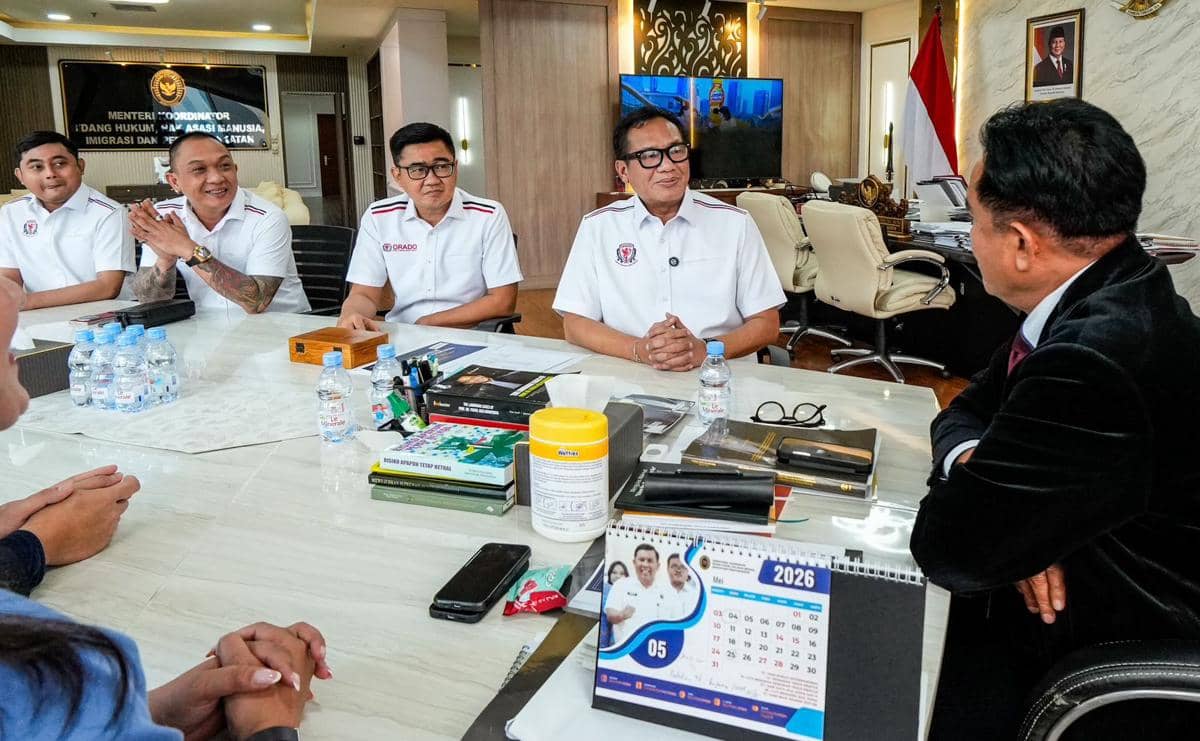 Beberapa pria mengenakan seragam putih ORADO duduk di meja rapat bersama seorang pejabat di kantor dengan bendera Indonesia di belakangnya.