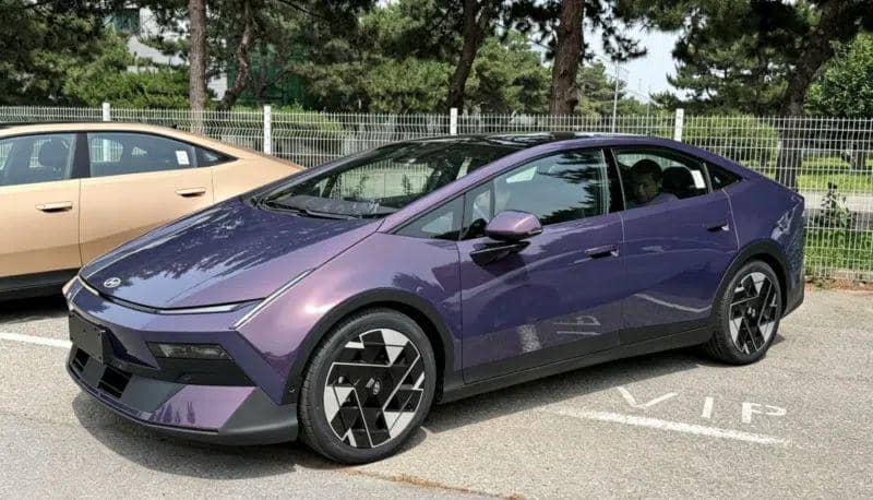 Mobil sedan listrik berwarna ungu metalik dengan desain futuristik diparkir di area luar dekat pagar dan pepohonan.