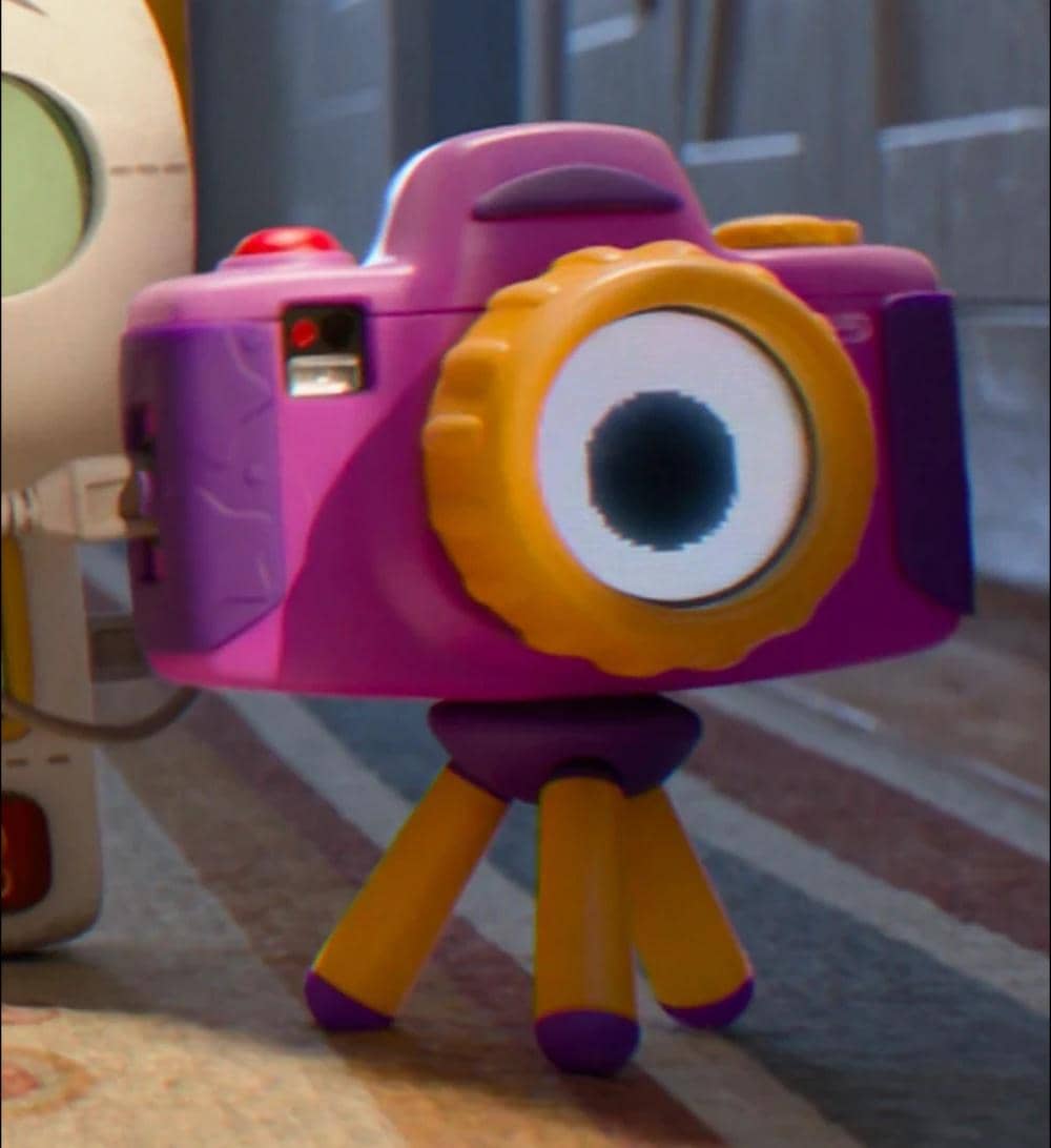 Kamera mainan berwarna ungu dan kuning dengan lensa berbentuk mata besar berdiri di atas lantai dalam cuplikan film animasi Toy Story 5.