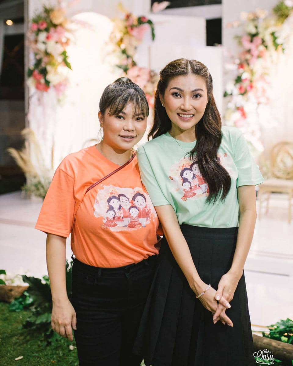 Dua perempuan berpose bersama mengenakan kaus bergambar karakter kartun dengan latar dekorasi bunga berwarna lembut.