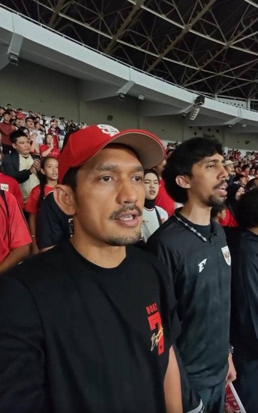 Dua pria mengenakan pakaian bertema timnas Indonesia berdiri di tribun stadion bersama penonton lain saat pertandingan Indonesia vs Oman.