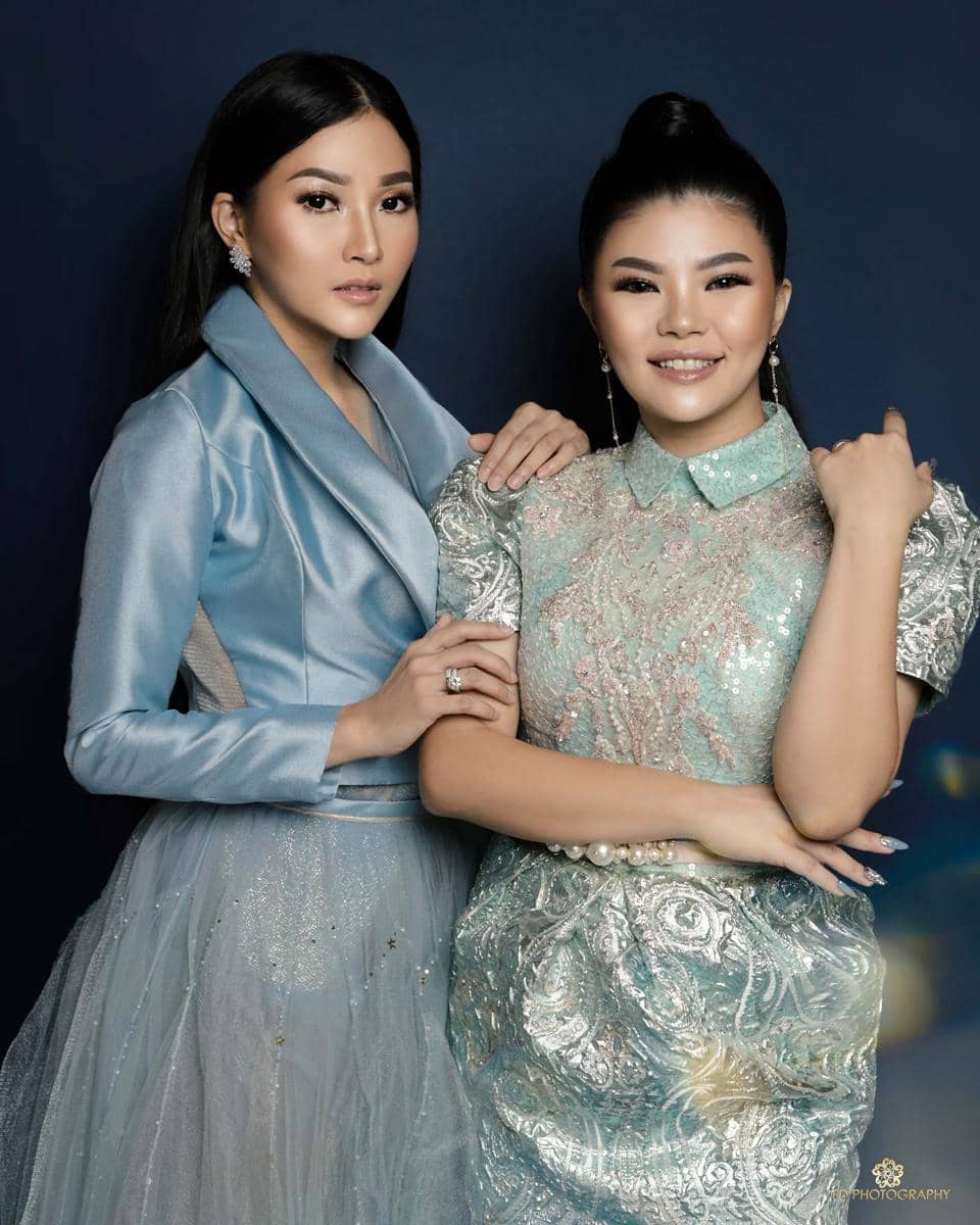 Dua perempuan berpose bersama mengenakan gaun elegan berwarna biru dan hijau pastel dengan latar belakang gelap.