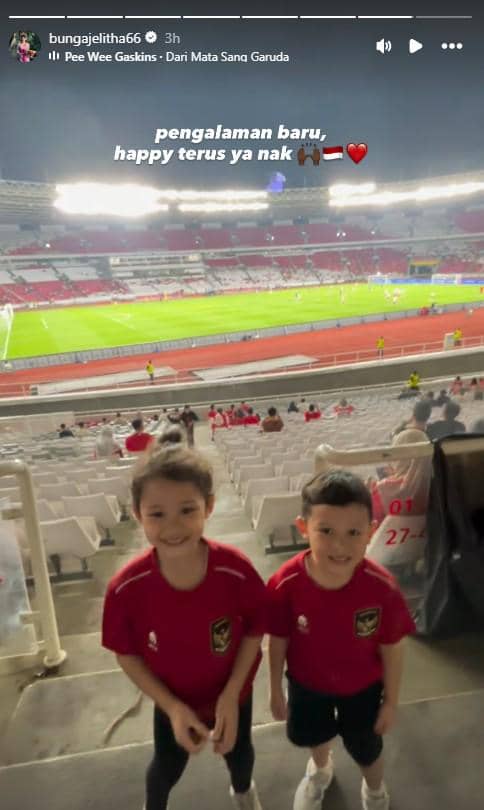 Dua anak mengenakan jersey timnas Indonesia berdiri di tribun stadion saat menonton pertandingan Indonesia melawan Oman.