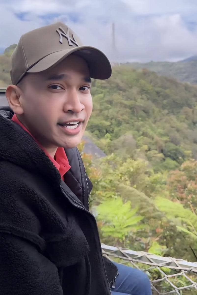 Seorang pria mengenakan jaket hitam dan topi cokelat duduk di area pegunungan hijau dengan latar langit berawan.
