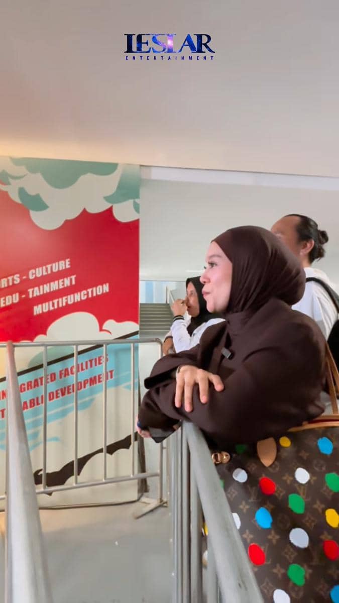 Seorang wanita berhijab cokelat berdiri di dekat pagar tribun sambil menonton pertandingan timnas Indonesia melawan Oman.