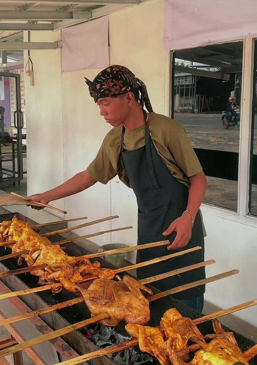 Seorang pria mengenakan apron hitam dan ikat kepala sedang memanggang ayam bakakak di atas bara api di Lembur Rasa Lampung.