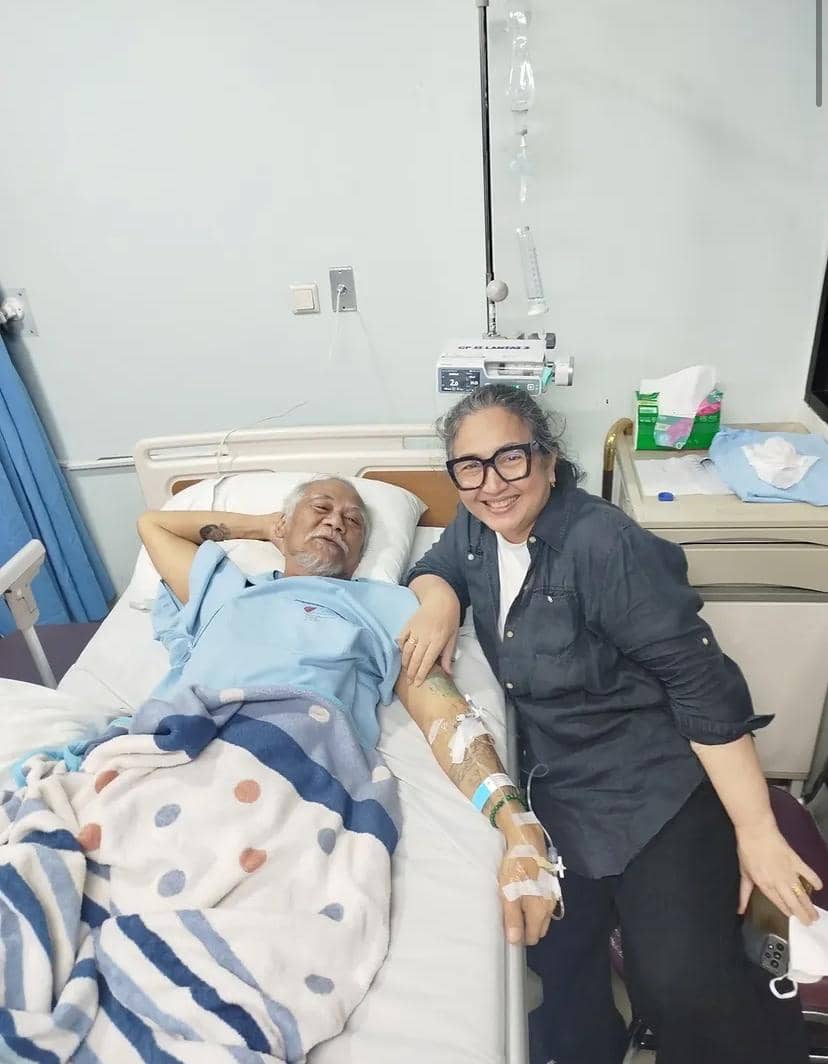 Seorang pria terbaring di ranjang rumah sakit dengan infus di lengannya, ditemani seorang wanita yang duduk di sampingnya sambil tersenyum.