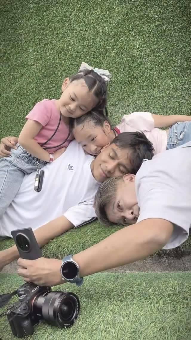 Seorang pria berbaring di rumput bersama tiga anak kecil yang tersenyum dan berpelukan sambil mengambil foto selfie dengan ponsel.