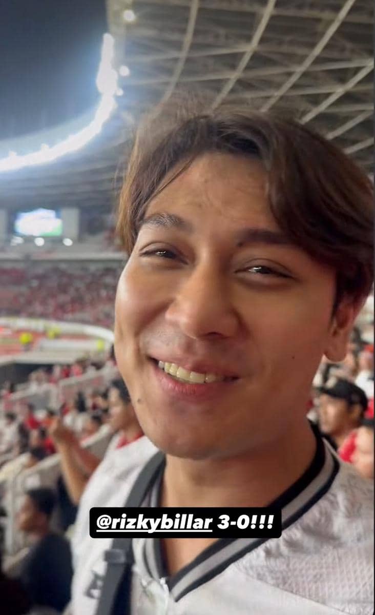 Seorang pria tersenyum di tribun stadion saat menonton pertandingan timnas Indonesia melawan Oman, dengan suasana ramai penonton di belakangnya.