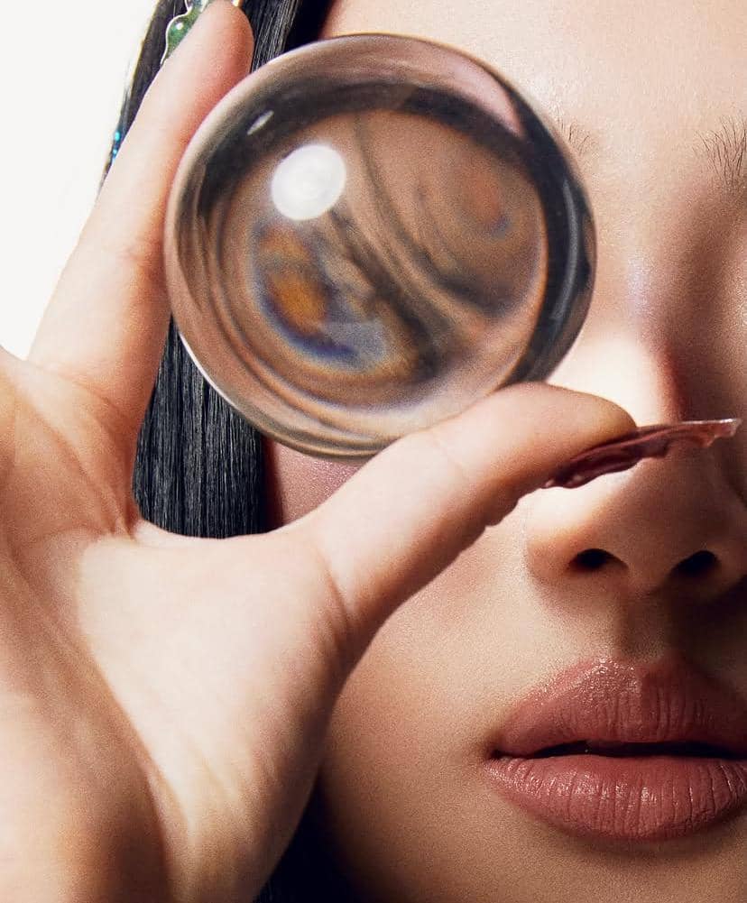 Seorang perempuan memegang bola kaca bening di depan wajahnya, memperlihatkan pantulan cahaya dan detail wajah secara artistik.