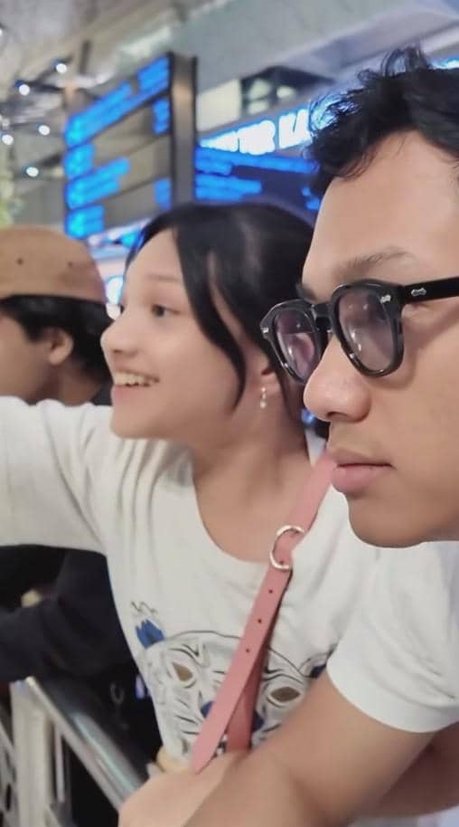 Seorang pria berkacamata dan seorang anak perempuan tersenyum sambil melihat ke arah tertentu di area bandara yang ramai.