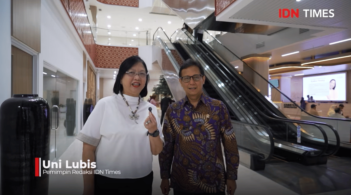 Dua orang berdiri di area lobi rumah sakit modern dengan eskalator di belakang, salah satunya memegang mikrofon kecil.