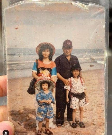 Sebuah foto keluarga lama di pantai memperlihatkan dua anak kecil dan dua orang dewasa mengenakan pakaian santai serta topi lebar.