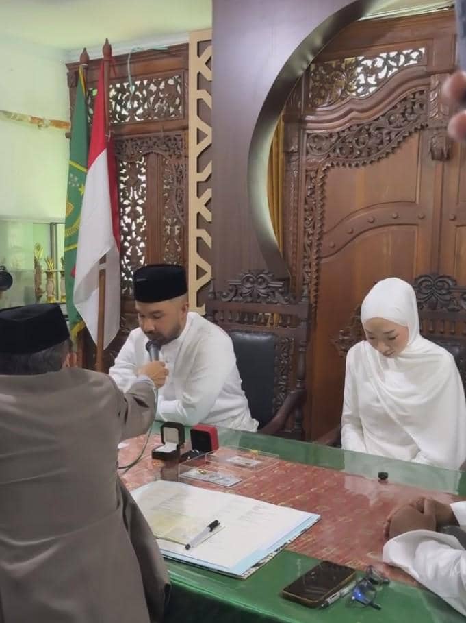 Pasangan mengenakan busana putih sedang melangsungkan akad nikah di KUA dengan penghulu di depan meja berisi dokumen dan cincin.