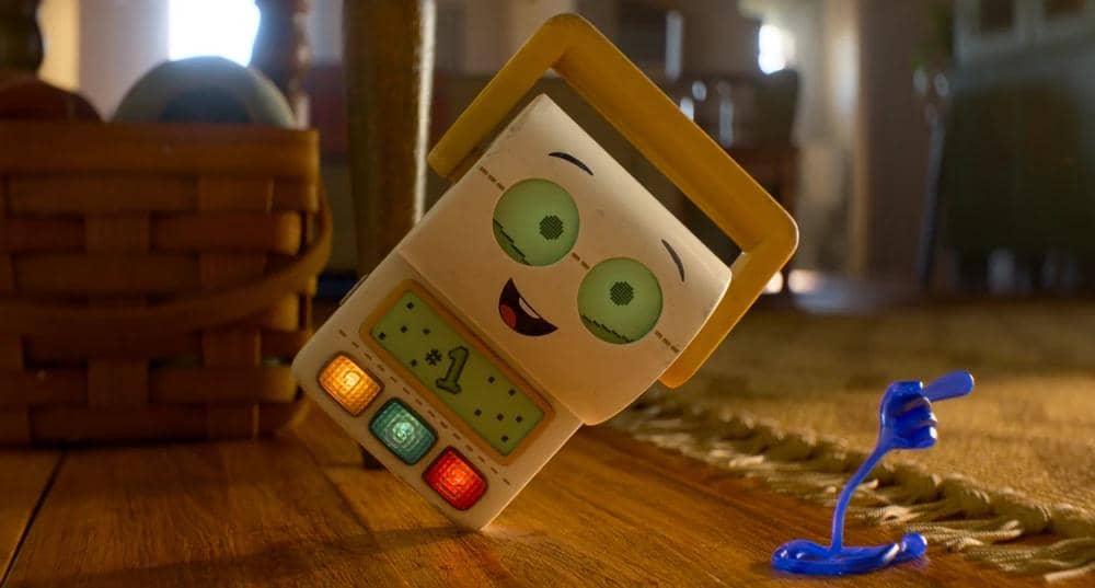 Karakter mainan baru berbentuk alat elektronik tersenyum di lantai kayu bersama mainan biru kecil dalam cuplikan film animasi Toy Story 5.