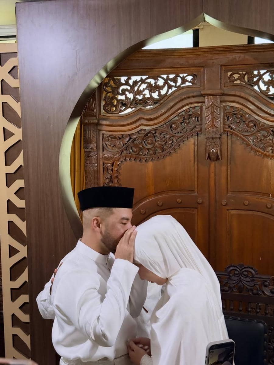 Seorang pria mengenakan baju koko putih dan peci hitam mencium kening wanita berhijab putih di depan ukiran kayu di KUA.