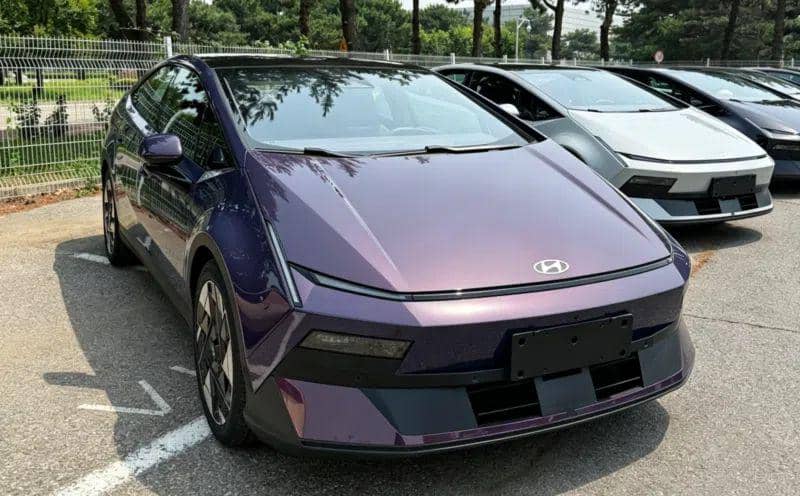 Sedan fastback berwarna ungu metalik dengan desain futuristik terparkir di area luar bersama beberapa mobil lain di belakangnya.