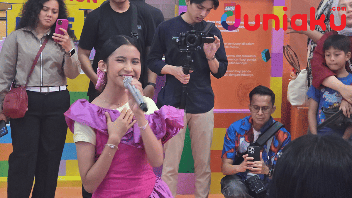 Seorang penyanyi muda mengenakan gaun ungu muda sedang bernyanyi dengan mikrofon di acara LEGO Playground di depan penonton.