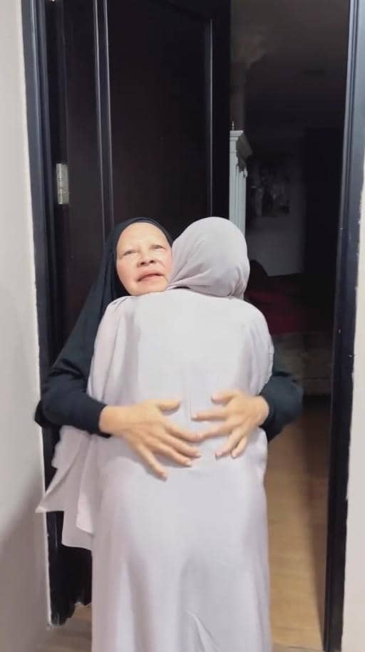 Dua perempuan berhijab berpelukan hangat di depan pintu rumah, salah satunya tampak terharu dengan mata tertutup.
