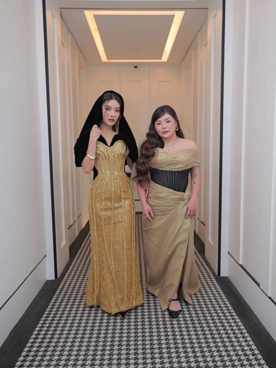 Dua wanita berpose di lorong dengan gaun elegan berwarna emas dan hitam, menampilkan gaya glamor dan percaya diri.
