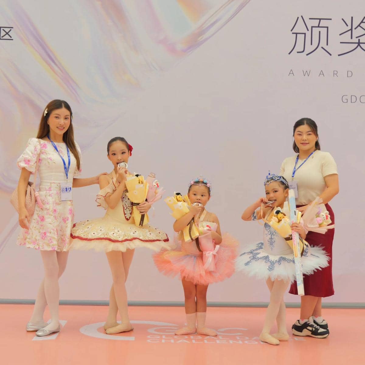 Dua wanita berpose bersama tiga anak kecil yang mengenakan kostum balet warna pastel sambil memegang buket bunga di atas panggung.