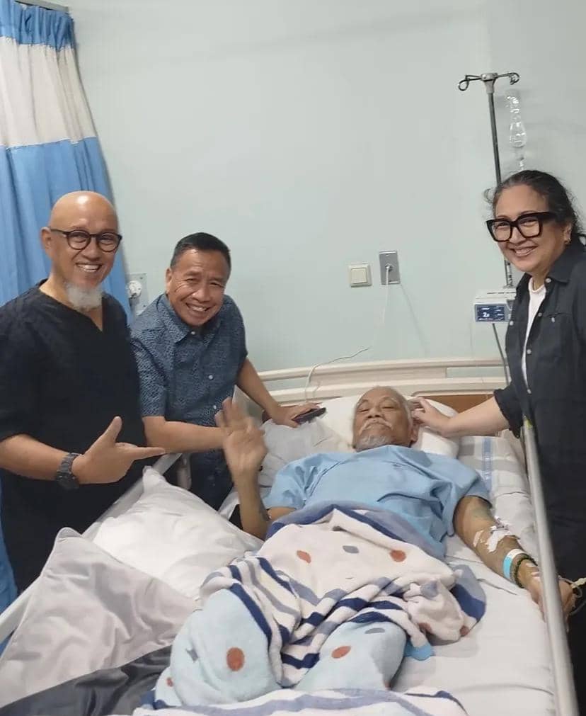 Seorang pria terbaring di ranjang rumah sakit mengenakan pakaian pasien, dikelilingi tiga orang yang tersenyum di sisinya.