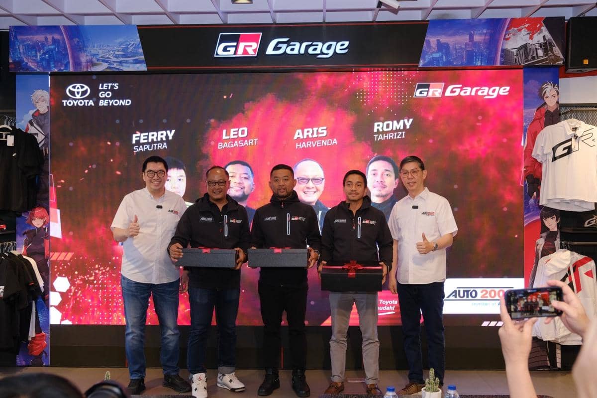 Para pembalap GRT berpose bersama tim Auto2000 di depan layar bertema GR Garage sambil memegang kotak hadiah.