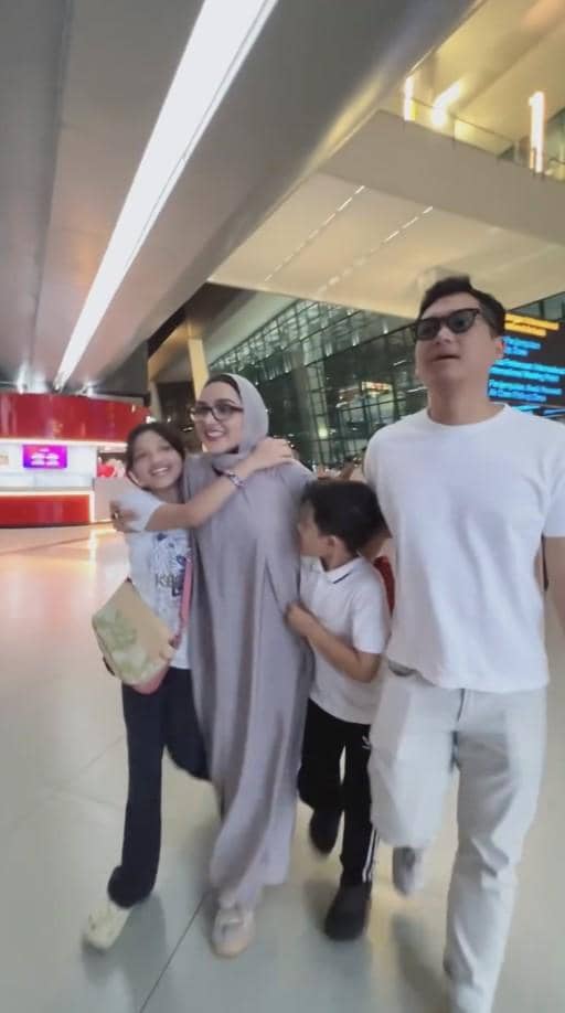Sebuah keluarga berjalan bersama di bandara dengan ekspresi bahagia, mengenakan pakaian santai dan membawa tas kecil.