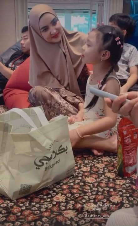 Seorang wanita berhijab cokelat tersenyum hangat kepada anak perempuan kecil yang duduk di depannya di ruang keluarga.