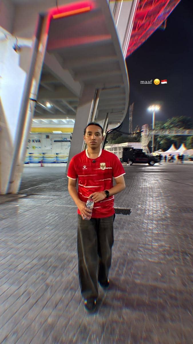 Seorang pria mengenakan jersey merah timnas Indonesia berdiri di area luar stadion pada malam hari sambil memegang botol air.
