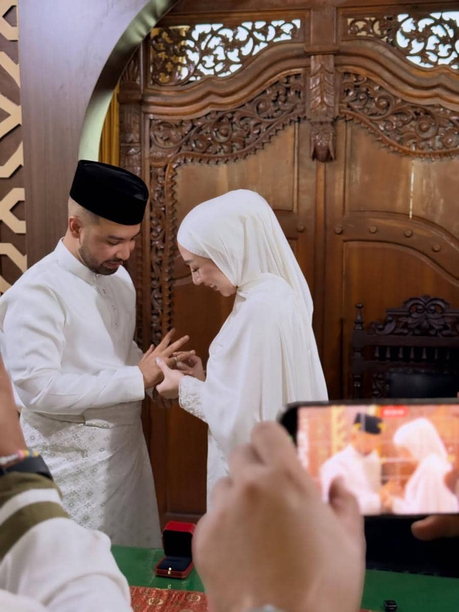 Pasangan mengenakan busana putih sedang bertukar cincin dalam prosesi pernikahan di KUA dengan latar ukiran kayu tradisional.