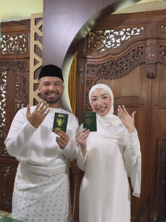 Sepasang pengantin berpakaian putih memperlihatkan buku nikah dan cincin pernikahan di Kantor Urusan Agama dengan latar kayu ukir.
