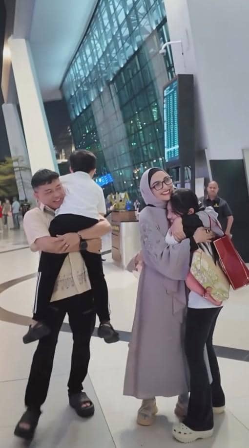 Sepasang suami istri bersama dua anak mereka berpelukan hangat di area kedatangan bandara setelah perjalanan ibadah haji.