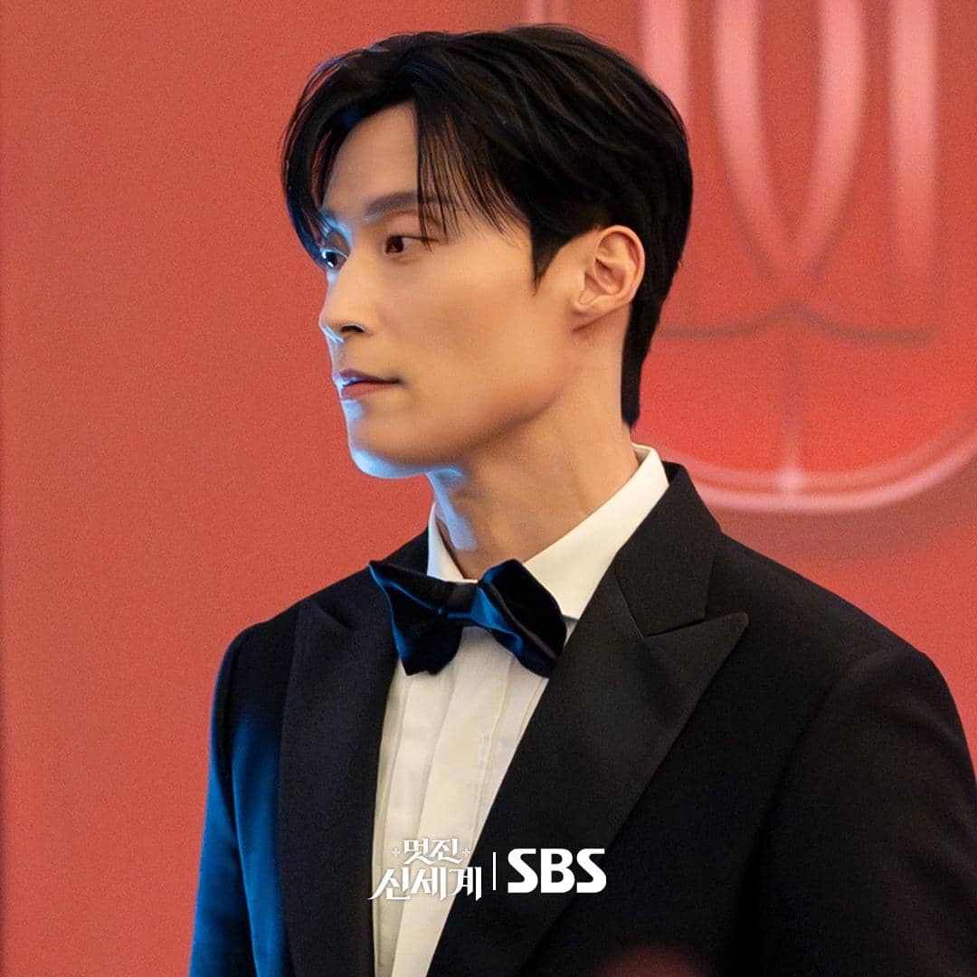 Seorang pria mengenakan setelan jas hitam dan dasi kupu-kupu berdiri di depan latar merah dalam cuplikan drama My Royal Nemesis.