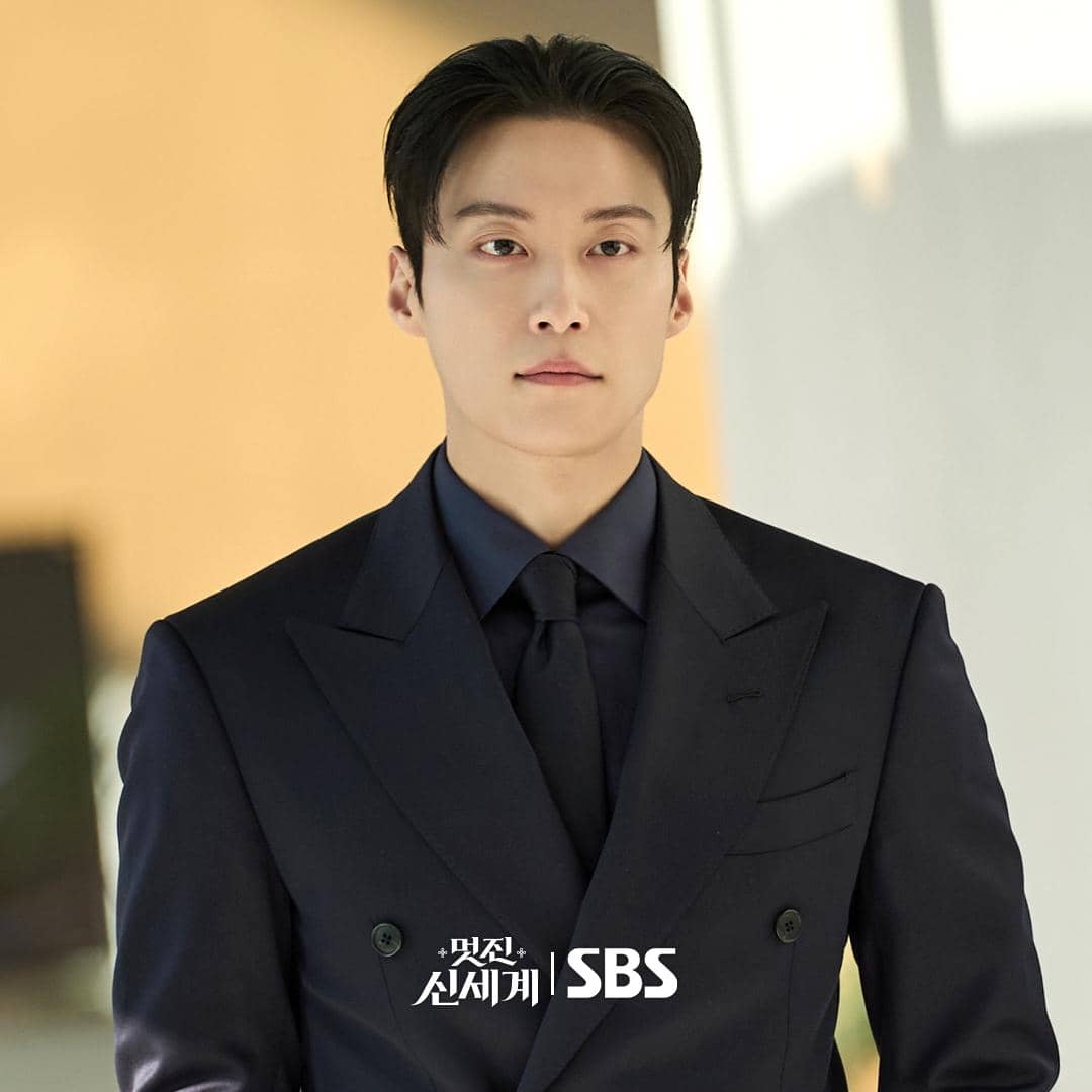 Seorang pria mengenakan setelan jas hitam berdasi gelap berdiri di dalam ruangan dengan pencahayaan hangat, cuplikan drama My Royal Nemesis.
