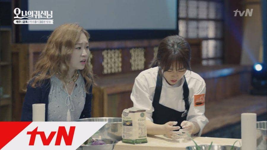 Dua wanita berada di dapur, salah satunya mengenakan seragam koki sedang menyiapkan adonan, sementara yang lain memperhatikannya.
