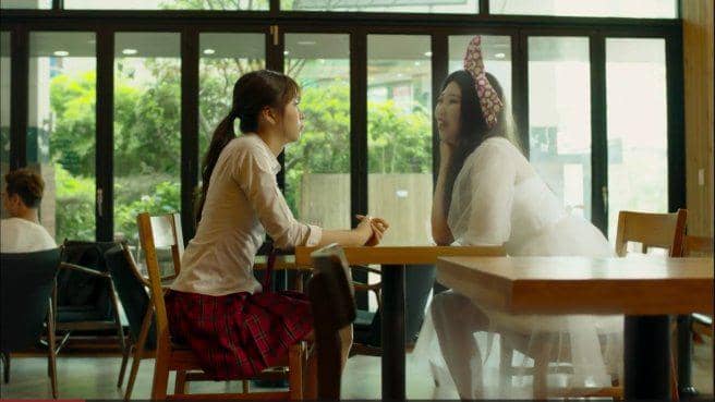 Dua perempuan duduk saling berhadapan di meja kafe, salah satunya mengenakan seragam sekolah dan yang lain berpakaian putih dengan pita besar di kepala.