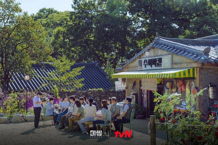 Beberapa orang duduk di luar bangunan tradisional Korea dengan atap biru, dikelilingi pepohonan dan suasana pedesaan yang tenang.