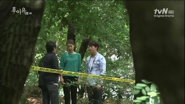 Tiga orang berdiri di area hutan yang dipasangi garis polisi kuning dalam cuplikan drama Who Are You dari tvN.